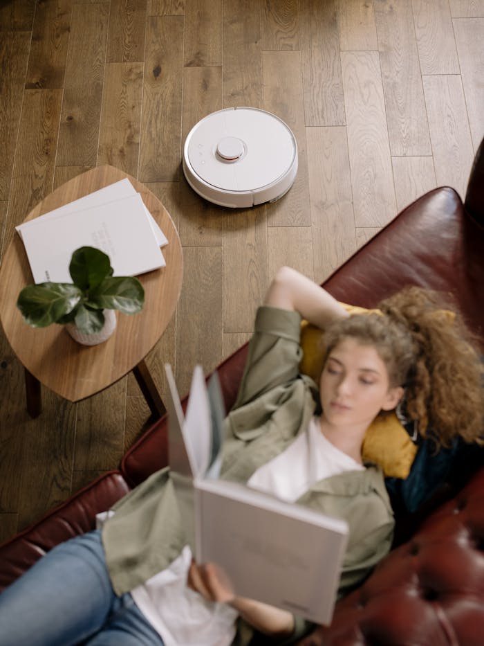 creative-02 Adult woman reading on a couch while a robot vacuum cleans the floor, epitomizing relaxation.